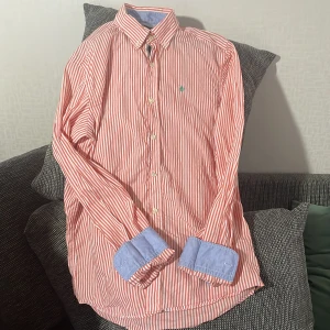 Randig skjorta från Morris, röd/vit M - Snygg skjorta från Morris i rödvitt randigt mönster med blå kontrast på insidan av krage och manschetter. Klassisk button down-krage, grönt broderat emblem på bröstet och vita knappar. Skjortan är långärmad och i skön bomull. Perfekt för en stilren look.