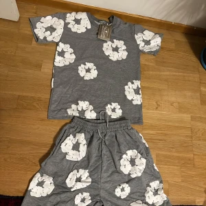 Grå t-shirt med blommor från Denim Tears - Grå t-shirt från Denim Tears med stora vita blommiga tryck över hela plagget. Klassisk passform med rund hals och korta ärmar. Mjuk bomullskvalitet som känns skön mot huden. Perfekt för dig som gillar streetwear med unik design.