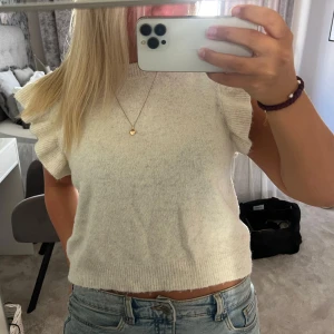 Vit stickad ärmlös tröja med volang - Superfin vit stickad tröja med volangdetaljer på axlarna. Modellen är croppad och ärmlös, perfekt till jeans eller kjol. Mjuk och skön, passar grymt för vår och sommar. Enkel att styla för en chill look.