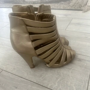 Guldiga stövletter med klack och remmar - Snygga guldiga stövletter med höga klackar och öppna partier med breda remmar. Skorna har öppen tå och dragkedja på sidan. Tillverkade i syntetmaterial med metallicfinish, perfekta för att sticka ut. Ger en edgy vibe till din outfit.