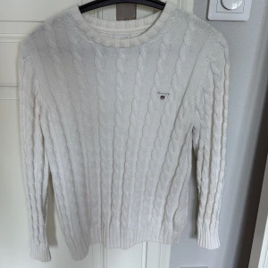 Vit kabelstickad tröja från Gant - Vit kabelstickad tröja från Gant med rund halsringning och diskret logga på bröstet. Tröjan är långärmad och har ribbade muddar vid ärmslut och nederkant. Perfekt för en clean och klassisk look.