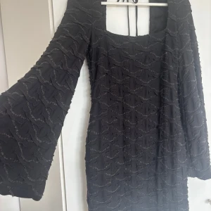 Svart strukturerad klänning med vida ärmar - Svart kortklänning med unik strukturerad yta och vågigt mönster. Klänningen har vida långärmar, fyrkantig ringning fram och djup, rundad rygg med knytband i nacken. Perfekt för dig som vill sticka ut med en cool och trendig look.