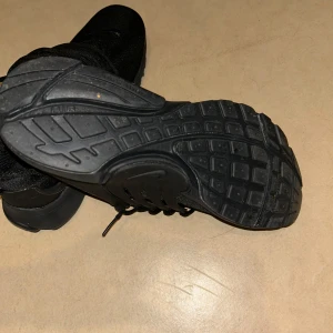 Svarta Nike sneakers med snörning - Snygga helsvarta Nike sneakers med klassisk snörning och robust sula. Skorna har en sportig look och är tillverkade i syntetmaterial med diskret Nike-logga på sidan. Perfekta för dig som gillar en clean och stilren stil.