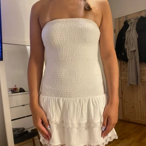 Vit axelbandslös kort klänning - Supersöt vit axelbandslös kort klänning med smockad överdel och volangkant nertill. Klänningen har spetsdetaljer längs fållen och är perfekt för varma dagar. Materialet känns lätt och luftigt, vilket gör den skön att bära.