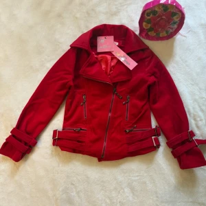 Röd bikerjacka med bältdetaljer - Snygg röd bikerjacka från Felomina med coola bältdetaljer vid ärmslut och midja. Jackan har flera dragkedjor framtill och en bred krage. Utsidan är mjuk och insidan har ett glansigt rött foder. Perfekt för dig som vill sticka ut med en färgstark look.