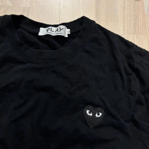 Svart Comme des Garçons Play t-shirt - Svart t-shirt från Comme des Garçons Play med klassisk rund hals och korta ärmar. På bröstet sitter det ikoniska hjärtat med ögon i svart. T-shirten är i mjuk bomull och har en clean, stilren vibe som passar till allt. Typ aldrig använd storlek L men sitter som en mindre M. Pris kan diskuteras 