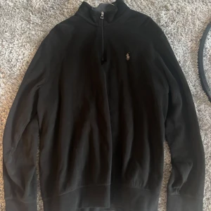 Svart half zip tröja från Ralph Lauren - Svart långärmad tröja från Ralph Lauren med half zip och klassisk broderad logga på bröstet. Hög krage och mjukt bomullsmaterial gör den både stilren och bekväm. Perfekt för dig som gillar enkel och clean stil.
