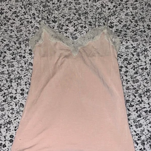 Ljusrosa linnetopp med spets från Oysho - Supersöt ljusrosa linnetopp från Oysho med tunna axelband och v-ringning dekorerad med vit spets. Toppen har en mjuk och flowy känsla, perfekt för lager-på-lager eller som statement piece. Enkel och feminin stil som passar till många looks.