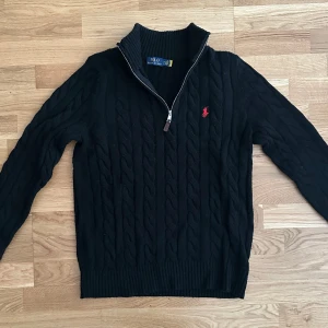 Svart kabelstickad tröja Polo Ralph Lauren - Svart kabelstickad tröja från Polo Ralph Lauren med röd broderad logga på bröstet. Tröjan har hög krage, half zip-dragkedja och långa ärmar. Klassisk och stilren modell i mjukt material, perfekt för kyligare dagar.