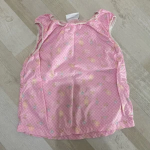 Rosa blommig ärmlös blus med spets - Supersöt rosa blus med små blommor och rutigt mönster. Ärmlös modell med volangkant och vit spets runt ärmhålen och halsen. Perfekt för dig som gillar gulliga och romantiska vibes.