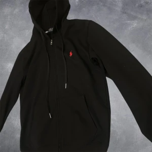 Svart hoodie från Polo Ralph Lauren - Svart hoodie från Polo Ralph Lauren med dragkedja, huva och snörning. Klassisk röd broderad logga på bröstet och stora fickor framtill. Tillverkad i mjuk bomull, perfekt för chill dagar eller när du vill ha en clean streetstyle.