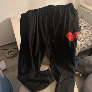 Svarta mjukisbyxor med hjärtdetalj - Svarta mjukisbyxor med resår i midjan och benslut, samt snörning i midjan. På ena benet finns ett rött trasigt hjärta tryckt som ger en cool och unik look. Perfekta för chill dagar eller streetwear-stil. Har även en matchande hoodie till