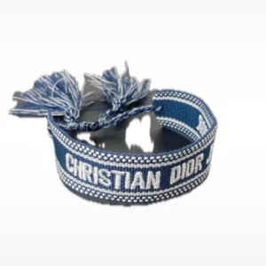 Blått armband från Christian Dior - Snyggt blått armband från Christian Dior med vit broderad logga och dekorativa tofsar i ändarna. Armbandet är justerbart med knytband och har en cool, avslappnad stil som passar perfekt till jeans eller som statement-accessoar.