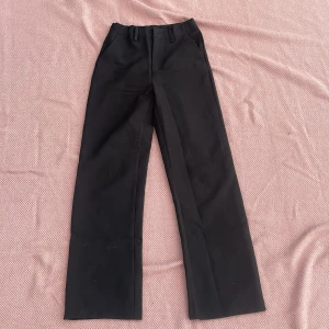 Svarta kostymbyxor XOXO Friends - Stilrena svarta kostymbyxor från XOXO Friends i storlek 152. Byxorna har raka ben, hällor för bälte och klassisk dragkedja med knapp. Perfekta för en clean och dressad look. Materialet känns mjukt och följsamt.