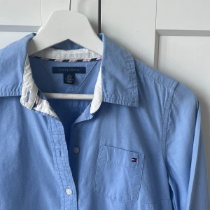 Blå skjorta från Tommy Hilfiger - Klassisk ljusblå skjorta från Tommy Hilfiger i bomull med vit detalj på insidan av kragen och diskret logga på bröstfickan. Skjortan har lång ärm, knappar hela vägen och en normal passform. Perfekt för en clean och stilren look.
