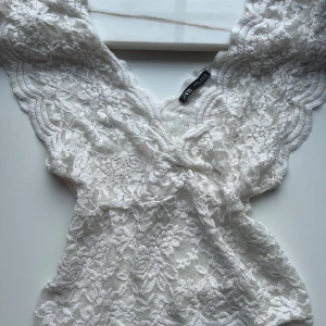 Vit spetsblus från Zara - Supersöt vit blus från Zara i genombruten spets med blommigt mönster. Djup v-ringning och ärmlös modell med vågiga kanter. Blusen är lätt och luftig, perfekt för varmare dagar.