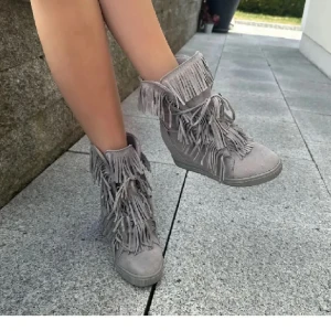 Grå mockaboots med fransar - Snygga grå boots i mocka med massor av fransar och snörning framtill. Skorna har en platt sula och rund tå, vilket gör dem både coola och bekväma. Perfekta för dig som vill sticka ut med en unik stil.
