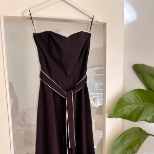 Svart jumpsuit med bälte - Elegant svart jumpsuit med axelbandslös design och vit kant längs överdelen. Klänningen har ett matchande svart bälte med vit piping i midjan som knyts framtill. Perfekt för dig som vill ha en stilren och klassisk look. Aldrig använd, strl 40