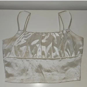 Champagnefärgad linnetopp från H&M - Snygg champagnefärgad linnetopp från H&M med tunna axelband och glansigt satinliknande material. Toppen har lätt rynkade detaljer framtill och är croppad i modellen. Perfekt för dig som vill ha en stilren och trendig look.