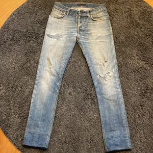 Ljusblå Nudie Jeans - Säljer ett par blå jeans med wash  från Nudie med slitningar och lagade hål på benen. 29/30. Klassisk femficksmodell med snygga detaljer på bakfickorna. De är väl använda men har mycket mer att ge. De är lagade på nudie butiken i Gbg och även uppsydda där,  så de nu är 30 på längden. 