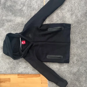 Svart Nike hoodiejacka med dragkedja - Svart hoodiejacka från Nike med hel dragkedja framtill och två fickor med dragkedja. Jackan har en klassisk huva och diskret Nike-logga på bröstet. Perfekt för chill dagar eller när du vill ha en sportig look.