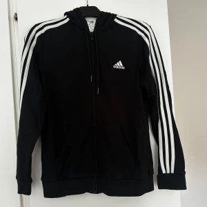 Svart Adidas hoodie med dragkedja - Svart hoodie från Adidas i storlek M med klassiska vita ränder längs ärmarna och Adidas-logga på bröstet. Koftan har huva med snörning, dragkedja framtill och två fickor. Tillverkad i mjuk polyesterblandning för skön passform.