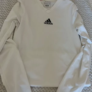 Vit långärmad Adidaströja  - Vit långärmad tröja från Adidas med svart logga på bröstet och V-ringning. Tillverkad i lätt och funktionell polyester med AEROREADY-teknologi som andas. Perfekt för träning eller sportiga outfits. Pris kan diskuteras🤍
