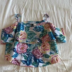 Blommig offshoulder topp med volang - Superfin offshoulder topp från Gina Tricot med tropiskt blommönster i rosa, blått, grönt och gult. Tunt tyg med volangdetalj och smala axelband. Perfekt för sommarens alla häng och picknickar!