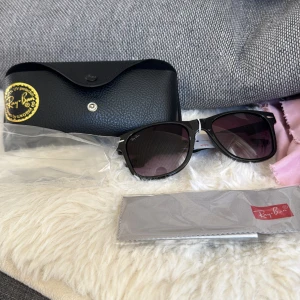 Svarta Ray-Ban Wayfarer solglasögon - Snygga svarta Ray-Ban Wayfarer solglasögon med mörka glas och klassisk form. Kommer med originalfodral och putsduk. Perfekta för att lyfta din stil och skydda ögonen mot solen. Solglasögon säljs billigt och helt nya pga att jag fick 70% hos synsam och köpte dessa. Och säljer nu för det är inte direkt min typ vill ha ett par bruna istället.