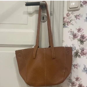 Brun axelväska  - Snygg brun axelväska i skinn med stilrena sömmar och både långa och handtag. Perfekt för att bära med sig det nödvändigaste. Väskan har en rymlig insida och en enkel design som passar till många tillfällen. Liknar väskan från Gina. Har inte använt den alls! 