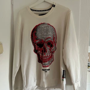 Vit sweatshirt med dödskalle Philipp Plein - Cool vit sweatshirt från Philipp Plein med stor dödskalle i svart och rött framtill. Tröjan har rund hals, långa ärmar och ribbade muddar. Märkesdetaljer på ärmen och halsen. Perfekt för dig som vill sticka ut med edgy streetstyle. Pris kan diskuteras ❤️❤️