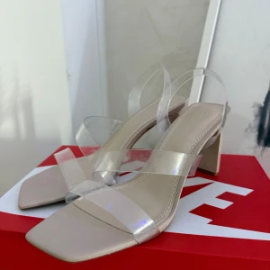 Beige sandaler med transparenta remmar - Stilrena sandaler i beige med fyrkantig tå och bred klack. Skorna har transparenta remmar i syntetmaterial som ger en modern och trendig look. Perfekta för dig som vill ha en clean och minimalistisk stil med en twist.