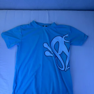 Blå Syna World T-shirt; (oanvänd) - (Finns att köpa tillsammans med matchade shorts!) (Pris kan diskuteras vid snabb affär) Snygg blå t-shirt med stort grafiskt tryck i svart och vitt som sträcker sig över både fram- och baksidan. Klassisk rund hals och korta ärmar. Perfekt för dig som gillar streetstyle och vill sticka ut med en cool design.