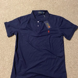 Marinblå pikétröja från Polo Ralph Lauren - Snygg marinblå pikétröja från Polo Ralph Lauren med klassisk krage, korta ärmar och knappar framtill. Ikoniska röda loggan broderad på bröstet. Tillverkad i mjuk bomull som känns skön mot huden. Perfekt för en clean och stilren look.