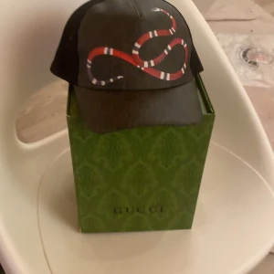 Gucci svart keps med ormtryck - Säljer en svart keps från Gucci med ikoniskt röd och vit ormtryck framtill. Kepsen har mesh på sidorna och justerbar rem baktill med röd och blå detalj. Kommer med originalbox och dustbag. Perfekt för dig som vill sticka ut med lyxig streetstyle.