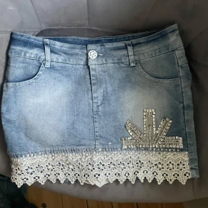 Jeanskjol med spets och strass - Snygg kort jeanskjol i ljusblå denim med dekorativa strasstenar och glittriga detaljer på ena sidan. Nederkanten har vit spets med blommönster som ger kjolen en unik och cool touch. Perfekt för dig som vill sticka ut lite extra.