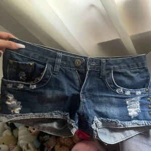 Blå jeansshorts med nitar och slitningar - Snygga blå jeansshorts med slitna detaljer, nitar och råa kanter. Ena bakfickan är i brun stickad stil med nitar, medan den andra har slitningar och nitar. Framsidan har slitna partier och dekorativa nitar vid fickorna. Perfekta för en edgy och cool look.