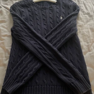 Mörkblå kabelstickad tröja Polo Ralph Lauren - Kabelstickad mörkblå tröja från Polo Ralph Lauren med klassisk rund halsringning och broderad logga på bröstet. Långärmad modell i mjuk bomull med ribbade muddar och nederkant. Perfekt för lager-på-lager och snygg till jeans.