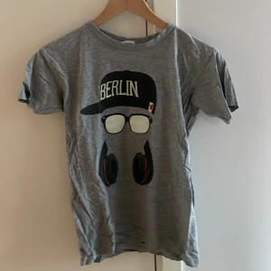 Grå t-shirt med Berlin-tryck B&C - Grå t-shirt från B&C Collection med coolt tryck av keps, glasögon och hörlurar samt texten 'Berlin' på framsidan. Klassisk passform och rund hals. Perfekt för dig som gillar streetstyle och vill sticka ut lite extra.