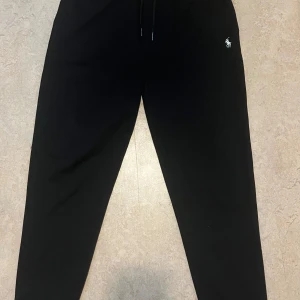 Svarta mjukisbyxor från Ralph Lauren - Svarta mjukisbyxor från Ralph Lauren med klassisk broderad logga på höften. Byxorna har dragsko i midjan och är tillverkade i mjukt material som är perfekt för chill dagar. Avsmalnande ben och enkel design gör dem lätta att matcha.