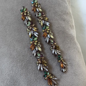 Långa glittriga örhängen med stenar - Superlånga örhängen med glittriga stenar i grönt, gult, orange, vitt och lila. Blommig design med många små stenar i olika former som ger en riktigt lyxig och färgstark look. Perfekta för dig som vill sticka ut och älskar statement-smycken.