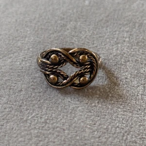 Exklusiv Kalevala ring i brons - Unik ring från Kalevala tillverkad i Finland med flätad design och fyra små guldfärgade kulor. Ringen har en antik vibe med detaljerade snirkliga mönster och är i en varm guldton. Perfekt för dig som gillar statement-smycken med retrostil. 