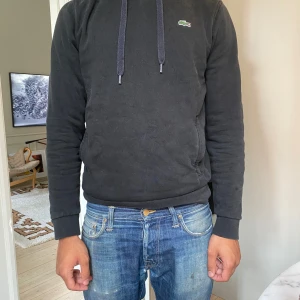 Svart hoodie från Lacoste - Svart hoodie från Lacoste med klassisk krokodil-logga på bröstet. Tröjan har huva med snörning, ribbade muddar och en avslappnad passform. Perfekt för dig som gillar en enkel och stilren look.