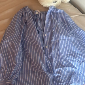 Blåvit randig skjorta från H&M - Snygg blå skjorta med vita ränder från H&M. Skjortan har klassisk krage, knäppning framtill och långa ärmar med manschett. Perfekt för en chill och stilren look. Tillverkad i mjuk bomull som känns skön mot huden.