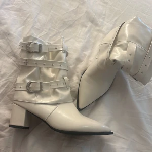 Beige boots med spännen och nitar - Snygga boots i glansigt cremevitt/beiget material med spetsig tå och blockklack. Dekorerade med tre remmar med silvriga spännen och små nitar för en edgy look. Aldrig använda 