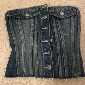 Jeans korsett-topp med knappar från Zara - Cool korsett-topp i blå denim med knappar framtill och rå kant nertill. Toppen har två fickor med lock och silverfärgade knappar. Snygga sömmar ger en markerad siluett. Perfekt för dig som vill sticka ut med en edgy look.