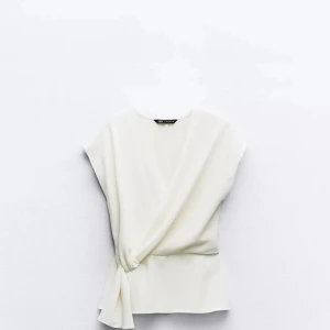 Vit omlottopp med knyt - Säljer en stilren vit omlottopp från Zara med knyt i sidan. Toppen har korta ärmar och en elegant v-ringning. Perfekt för dig som gillar en enkel men snygg look.