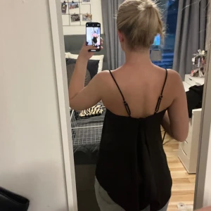 Svart linnetopp med tunna axelband - Säljer en svart linnetopp med tunna, justerbara axelband och rak halsringning. Toppen har en lös passform och är perfekt för varma dagar eller att styla lager på lager. Enkel och stilren design som funkar till mycket. Knyt själv i ryggen för att välja hur öppen rygg du vill ha. Från ginatricot.