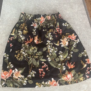 Svart blommig kortkjol med resår - Supersöt svart kortkjol med färgglatt blommönster och elastisk midja. Kjolen har gröna blad, orange och vita blommor samt små fåglar i printet. Perfekt för dig som vill sticka ut med en somrig vibe.