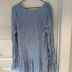 Ljusblå blommig klänning från NA-KD - Supersöt ljusblå kortklänning från NA-KD med broderade blommor över hela tyget. Klänningen har långa ärmar med volangdetaljer och en djup rygg. Perfekt för vår och sommar när du vill känna dig extra fin.